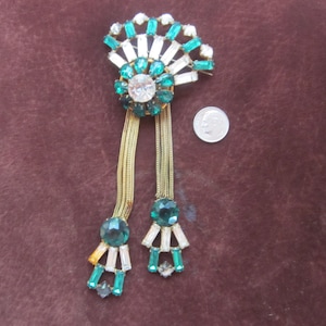 May include: A vintage green and clear rhinestone brooch with dangling chains. The brooch is shaped like a fan and has a large, round rhinestone in the center.