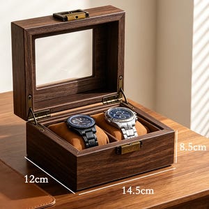 Op de afbeelding: Een donkerbruine horlogedoos met 2 sleuven en een doorzichtig glazen bovenkant, met twee horloges. De doos is 14,5 cm lang, 12 cm breed en 8,5 cm hoog. De tekst "2-slot watch box" staat bovenaan.