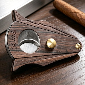 May include: A cigar cutter with a dark brown wood grain finish and a silver circular blade opening. The tool features a gold-toned metal knob and a small screw on the side. The word Feilide is engraved in a cursive script on the wooden surface.