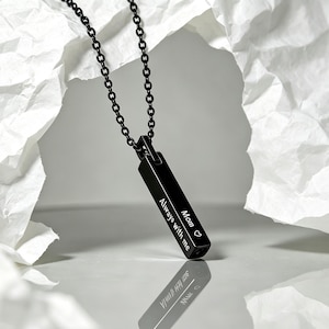 May include: A black metal bar pendant necklace featuring a delicate chain and a rectangular charm engraved with the text "Mom" followed by a small heart symbol and the phrase "Always with me" in a clean, white, sans-serif font style.