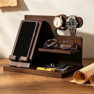 K&ouml;nnte beinhalten: Ein dunkelbrauner Schreibtisch-Organizer aus Holz mit einem schr&auml;gen St&auml;nder f&uuml;r ein Smartphone, einer horizontalen Ablage f&uuml;r eine Brille und einer oberen Leiste f&uuml;r zwei Armbanduhren. Die Basis enth&auml;lt ein vertieftes Fach f&uuml;r einen Schl&uuml;sselbund.