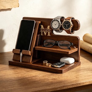 K&ouml;nnte beinhalten: Eine Ladestation aus dunklem Holz mit einem Handyhalter inklusive Ladekabel, einer Ablage f&uuml;r eine Brille und einer horizontalen Stange f&uuml;r zwei Armbanduhren. Das Holz ist mit dem Buchstaben M, dem Namen MALACHI und der r&ouml;mischen Zahl IV graviert.