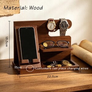K&ouml;nnte beinhalten: Eine dunkelbraune Holz-Ladestation mit einer H&ouml;he von 18,5 cm, einer Breite von 22,5 cm und einer Tiefe von 15 cm. Sie verf&uuml;gt &uuml;ber einen Handyhalter, eine Ablage f&uuml;r Brillen, zwei Haken f&uuml;r Uhren und ein Fach f&uuml;r Schl&uuml;ssel und M&uuml;nzen.