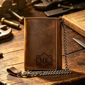 May include: A brown leather trifold wallet featuring a dark engraved diamond-shaped emblem with the letters MK in the center. The wallet includes a metal chain attached to a leather loop with a snap button, showcasing visible stitching along the edges.
