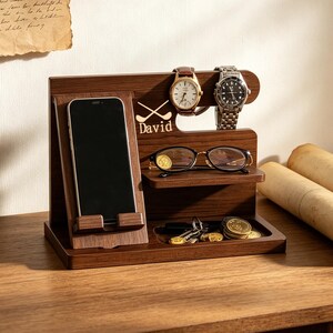 K&ouml;nnte beinhalten: Ein dunkelbrauner Schreibtisch-Organizer aus Holz mit einem vertikalen Smartphone-St&auml;nder, einer Ablage f&uuml;r eine Brille, zwei Halterungen f&uuml;r Armbanduhren und einem kleinen Fach f&uuml;r Schl&uuml;ssel und M&uuml;nzen. Das Holz ist mit Golfschl&auml;gern und David graviert.