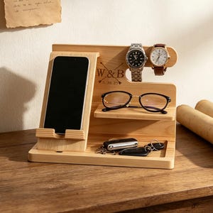 K&ouml;nnte beinhalten: Eine Dockingstation aus hellem Holz mit einem Smartphone-St&auml;nder, einer Ablage f&uuml;r eine Brille und Haken f&uuml;r zwei Armbanduhren. In der Mitte ist der Text W & B und 5. 30. 25 eingraviert. Ein Schl&uuml;sselbund liegt in der Ablageschale des Organizers.