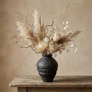 May include: A black textured vase filled with dried pampas grass, protea flowers, and delicate branches. The arrangement sits on a rustic wooden table, creating a natural and elegant display.