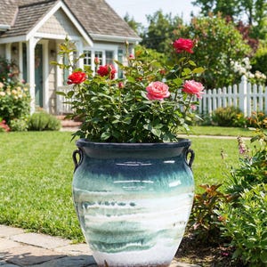 May include: A large, glazed ceramic planter with a blue and green ombre effect, filled with a rose bush bearing red and pink blooms. The planter has two curved handles and sits on a brick walkway in a garden setting.