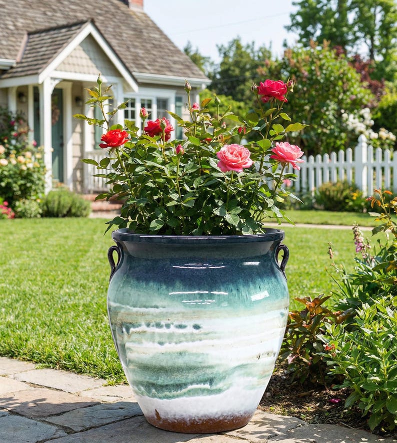 May include: A large, glazed ceramic planter with a blue and green ombre effect, filled with a rose bush bearing red and pink blooms. The planter has two curved handles and sits on a brick walkway in a garden setting.