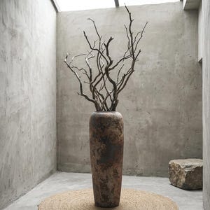 Könnte beinhalten: Eine hohe, strukturierte, braune Vase gefüllt mit getrockneten Ästen. Die Vase hat ein rustikales, gealtertes Aussehen und steht auf einer runden, gewebten Matte. Der Hintergrund ist ein minimalistischer Raum mit Betonwänden und natürlichem Licht.