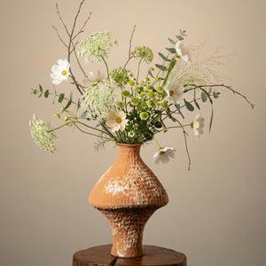 Puede incluir: Un jarrón de terracota texturizado contiene un vibrante arreglo de cosmos blancos y amarillos, encaje de la reina Ana y vegetación. El jarrón se encuentra sobre una superficie de madera, creando una estética natural. Ideal para la decoración del hogar.