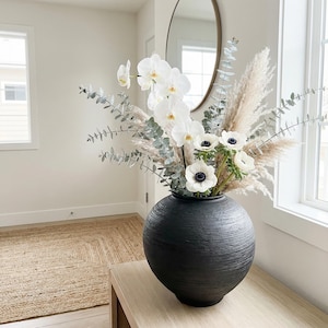 Könnte beinhalten: Eine schwarze, strukturierte Keramikvase enthält ein Blumenarrangement. Weiße Orchideen, Anemonen, Pampasgras und Eukalyptuszweige bilden eine natürliche, elegante Präsentation. Die Vase steht auf einem hellen Holztisch, im Hintergrund ein Teppich in neutralen Tönen.