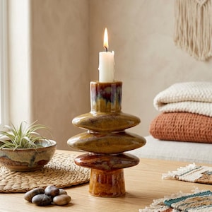 May include: A glazed ceramic candle holder with a lit white candle. The holder has a brown and yellow glaze. A small bowl with a plant and a woven placemat are nearby. Decorative items are on a wooden surface.