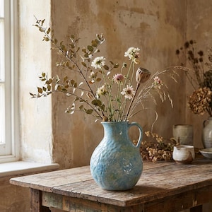 Puede incluir: Un jarrón de cerámica azul claro con una superficie texturizada, lleno de una variedad de flores secas y follaje. El jarrón está sobre una mesa de madera rústica, con una ventana y una pared de tonos neutros en el fondo.