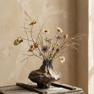 May include: A decorative floral arrangement in a textured, dark brown vase. The bouquet includes dried grasses, branches, and delicate flowers in shades of cream, purple, and tan. The vase sits on a rustic wooden surface.