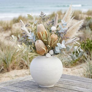 Puede incluir: Un jarrón de cerámica blanco texturizado contiene un arreglo floral de temática costera. El ramo presenta flores de banksia secas, hierba de la Pampa, eucalipto y madera flotante. Se incluyen pequeñas conchas marinas.