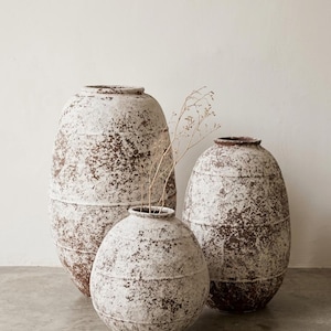 Könnte beinhalten: Drei verwitterte, cremefarbene Keramikvasen mit braunen Akzenten. Die Vasen variieren in Größe und Form, mit einer großen, länglichen Vase links, einer kleineren runden Vase in der Mitte und einer mittelgroßen Vase rechts. Getrocknete Stiele befinden sich in der mittleren Vase.