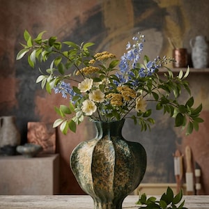 May include: A textured, green and gold vase holding a colourful floral arrangement. The bouquet features yellow, white, and blue flowers, with green leaves. The vase is placed on a wooden table. The vase is likely made of ceramic.