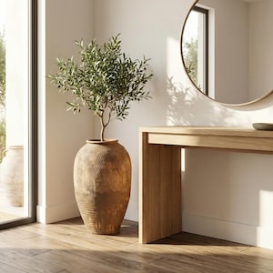 Könnte beinhalten: Ein großer, strukturierter, brauner Keramiktopf mit einem kleinen Olivenbaum. Der Topf ist hoch und zylindrisch mit vertikalen, geschnitzten Linien. Der Baum hat grüne Blätter und kleine schwarze Oliven. Eine Holzkonsolentisch und ein runder Spiegel sind im Hintergrund.