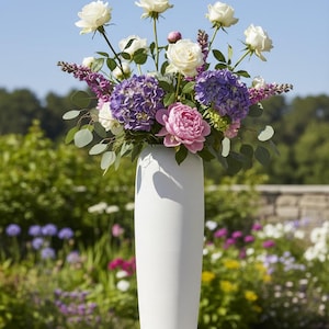 May include: A white vase holding a colourful floral display. The arrangement features white roses, purple hydrangeas, pink peonies, and green foliage. The vase is tall and cylindrical with a textured finish, set against a garden background.