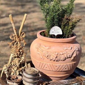 May include: A terracotta plant pot with a detailed relief design of a garland and ribbons. The pot contains a small evergreen plant and a white plant marker. Beside the pot are small, rustic metal bowls and decorative elements.