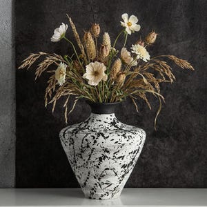 Può includere: Un vaso bianco con un design a spruzzi di vernice nera, contenente un bouquet di fiori ed erbe secche. Il vaso ha un bordo nero e una forma svasata. I fiori sono beige e bianchi, creando un'estetica naturale e rustica.