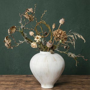 Puede incluir: Un jarrón blanco y redondo con un acabado texturizado contiene un arreglo floral seco. El arreglo incluye flores secas, cápsulas de semillas y ramas con hojas y musgo. El fondo es verde oscuro.