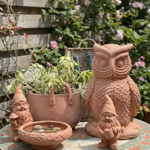 Puede incluir: Una estatua de búho de terracota y gnomos de jardín se exhiben sobre una mesa con mosaicos. También hay una maceta de terracota con vegetación y un abrevadero para pájaros. La escena está ambientada en un jardín con una valla de madera y rosales.