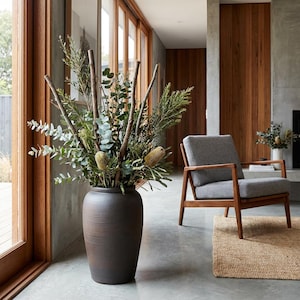 May include: A tall, dark brown textured ceramic vase filled with dried greenery and branches. The vase is in a modern living space with a grey armchair and wooden features. The vase is likely around 75 cm tall.