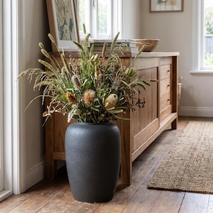 May include: A large, dark grey textured vase filled with dried flowers and grasses. The vase sits on a wooden floor in front of a wooden cabinet. The arrangement includes various shades of green, brown, and beige.