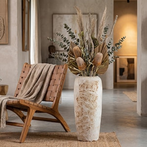 May include: A tall, textured white vase filled with dried flowers and foliage, including eucalyptus and large, brown blooms. A woven leather chair with a tan blanket sits to the left. The scene is bathed in warm, natural light, creating a cozy, inviting atmosphere.
