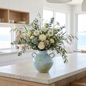 May include: A blue vase with a floral arrangement of white flowers and green foliage. The vase has two handles and sits on a light-coloured countertop. The room is bright and airy, with a view of the sea visible through the windows.