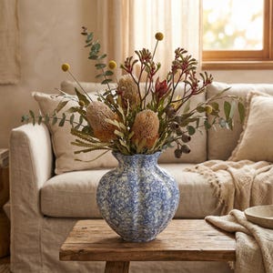May include: A blue and white speckled ceramic vase filled with dried flowers, including large tan banksia blooms, eucalyptus, and other greenery. The vase sits on a rustic wooden table in a living room setting.