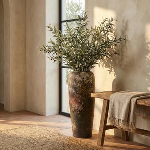 Può includere: Un vaso alto in ceramica anticata pieno di rami di ulivo. Il vaso ha una finitura screziata marrone e rossa. I rami di ulivo hanno foglie verdi e piccole olive nere. A destra c'è una panca di legno con una coperta beige.
