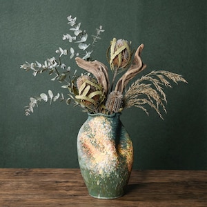 May include: A decorative vase with a green and yellow speckled design, filled with dried flowers, eucalyptus, and driftwood. The arrangement sits on a wooden surface against a dark green backdrop. A natural, rustic home decor item.