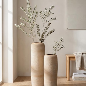 Könnte beinhalten: Zwei beige strukturierte Keramikvasen unterschiedlicher Höhe, jede mit belaubten grünen Zweigen. Die größere Vase ist etwa 91 cm hoch, die kleinere etwa 61 cm. Die Vasen stehen auf einem hellen Holzboden.