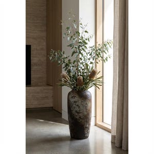 May include: A large, aged-effect vase filled with foliage and dried flowers. The vase has a dark brown and grey mottled finish. The arrangement includes eucalyptus branches and several tan-coloured, cone-shaped blooms. The vase is near a window with neutral-toned curtains.