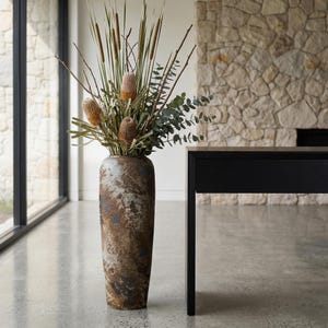 May include: A tall, textured vase in shades of brown and gray, filled with dried flowers and greenery. The vase has a rustic, weathered appearance and is placed in a modern interior setting. The arrangement includes cattails, eucalyptus, and other decorative elements.