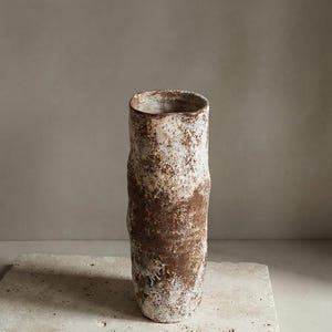 Puede incluir: Un jarrón cilíndrico de cerámica alto con una forma orgánica e irregular. La superficie presenta un acabado texturizado y moteado con una mezcla de tonos marrón tierra y blanco crema, creando un aspecto rústico y desgastado con variaciones.
