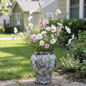 Puede incluir: Un jarrón grande y texturizado lleno de un vibrante arreglo de flores de cosmos y dedaleras. El jarrón tiene un patrón moteado en blanco y negro con detalles en verde. Las flores son blancas y rosas.
