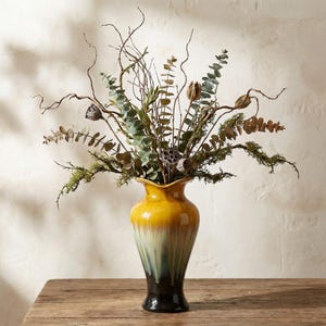 May include: A decorative vase with a yellow, green, and black ombre design, filled with dried branches, eucalyptus leaves, and moss. The vase sits on a wooden surface, creating a natural, earthy aesthetic.
