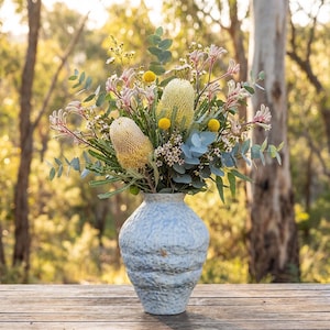 May include: A light blue ceramic vase filled with a vibrant floral arrangement. The bouquet includes yellow, pink, and white flowers, along with green eucalyptus leaves. The vase sits on a wooden surface, with a blurred forest background.