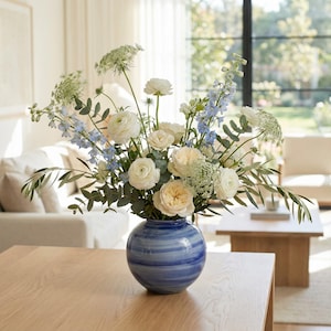 Pode incluir: Um arranjo floral num vaso listrado azul e branco. O buquê apresenta rosas brancas e creme, delfínios azuis e folhagem verde. O vaso está sobre uma mesa de madeira clara.