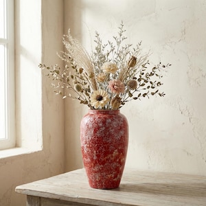 May include: A tall, red speckled vase filled with dried flowers and grasses. The vase has a textured surface and a wide base, tapering towards the top. The arrangement includes beige, pink, and brown floral elements, creating a natural, rustic aesthetic.