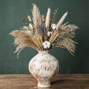 Puede incluir: Un arreglo decorativo de flores secas en un jarrón blanco y marrón. El ramo incluye hierba de la pampa, trigo, lavanda, algodón y eucalipto. El jarrón tiene forma redonda y un acabado pintado envejecido. El fondo es una pared verde oscuro.