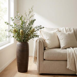 May include: A tall, textured brown vase filled with green and yellow floral arrangements. The vase is positioned next to a beige sofa with cream-colored pillows and a fringed throw blanket. A window is visible in the background.