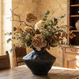 May include: A large, black, textured vase filled with a dried floral arrangement. The arrangement includes dried hydrangeas, eucalyptus, and various seed pods and branches. The vase sits on a wooden table in a room with a stone wall and wooden furniture.