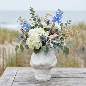 Puede incluir: Un arreglo floral de temática costera en un jarrón blanco. El ramo incluye hortensias blancas, delfinios azules y hierbas secas. La madera flotante y las conchas marinas se suman a la estética playera. El jarrón tiene un aspecto texturizado y marmolado.