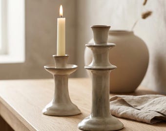 May include: Two ceramic candle holders in a neutral color palette. One candle holder has a lit, cream-colored candle. A beige vase and a linen cloth are in the background. The candle holders are on a wooden surface.