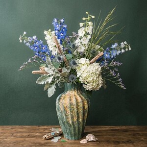 May include: A vase of flowers in shades of blue, white, and green. The vase is green and gold. The arrangement includes delphiniums, hydrangeas, and thistle. Driftwood and shells are on the wooden table.